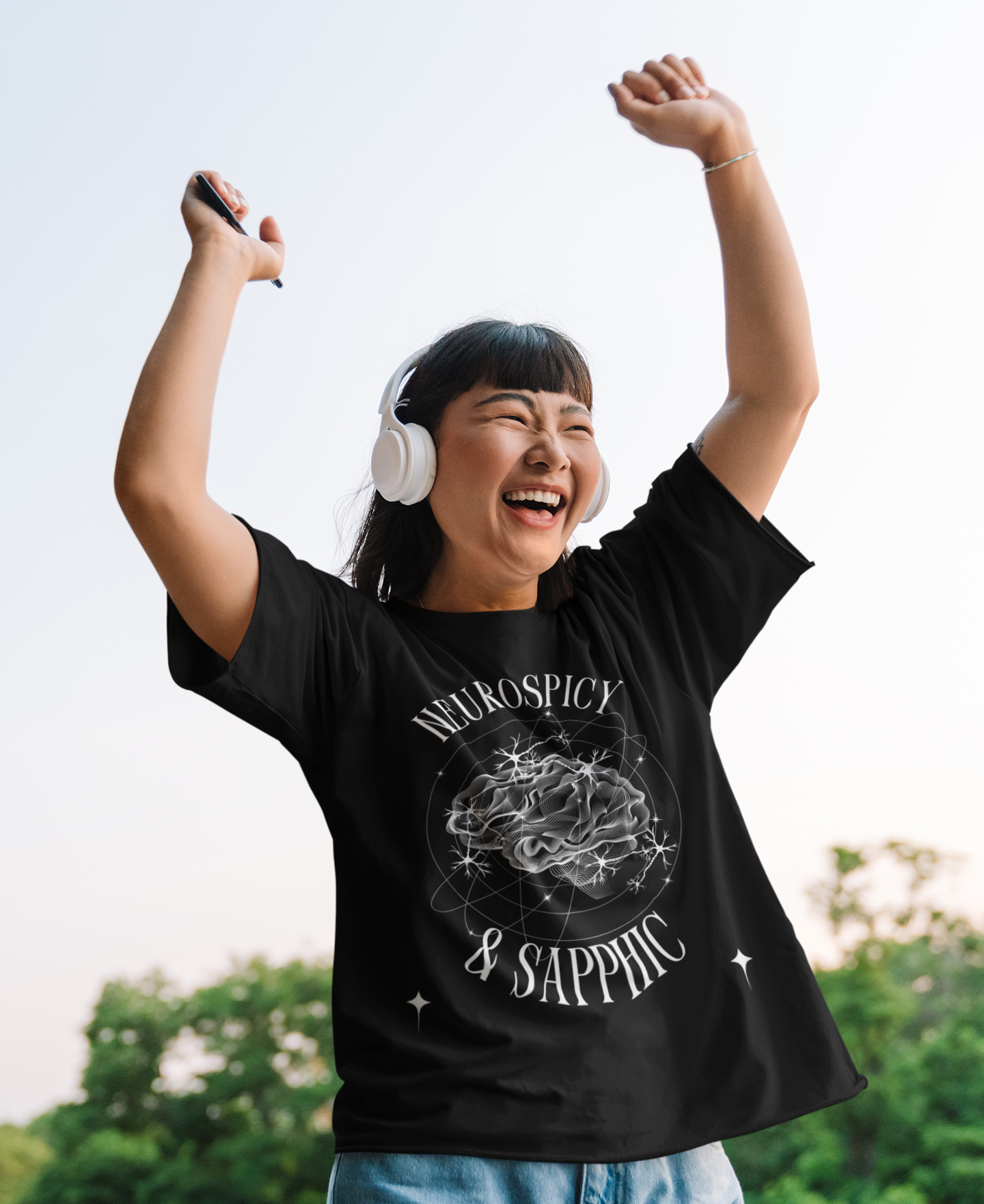 Asian woman cheering with white headphones and an oversize black t-shirt that says neurospicy and sapphic with streetwear inspired design for neurodivergent pride