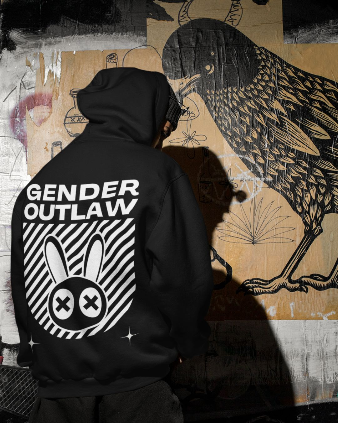 Urban scene with someone in a black hoodie that says Gender Outlaw across the back with the Mad Rabbit streetwear-inspired design and two stars for Trans Pride, Enby Pride, Nonbinary Pride, Genderdiverse Pride