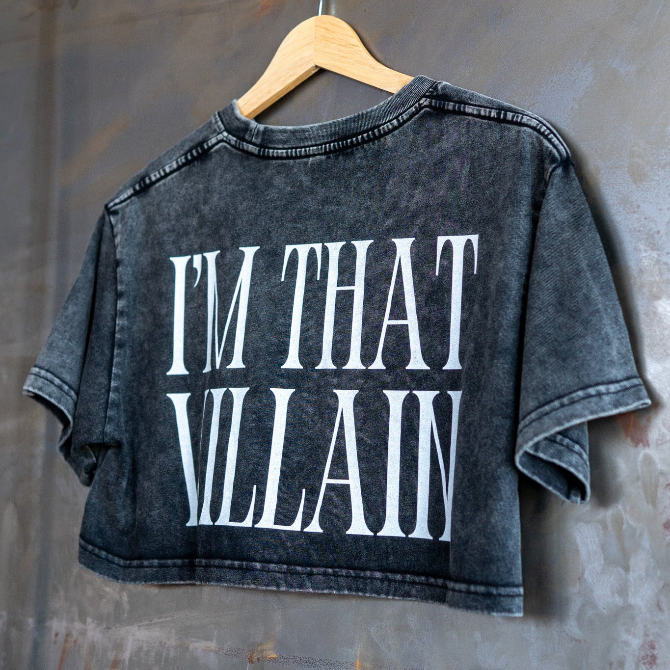 Black acid wash crop top with 'I'm That Villain' text on a hanger against a textured wall.