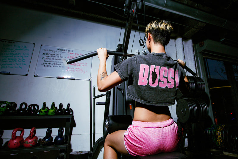 non-binary Person at the gym exercising and wearing a snow wash crop top with 'BUSSY' on the back with a Cat for BUSSY Cat Club series by Nova Edge Threads