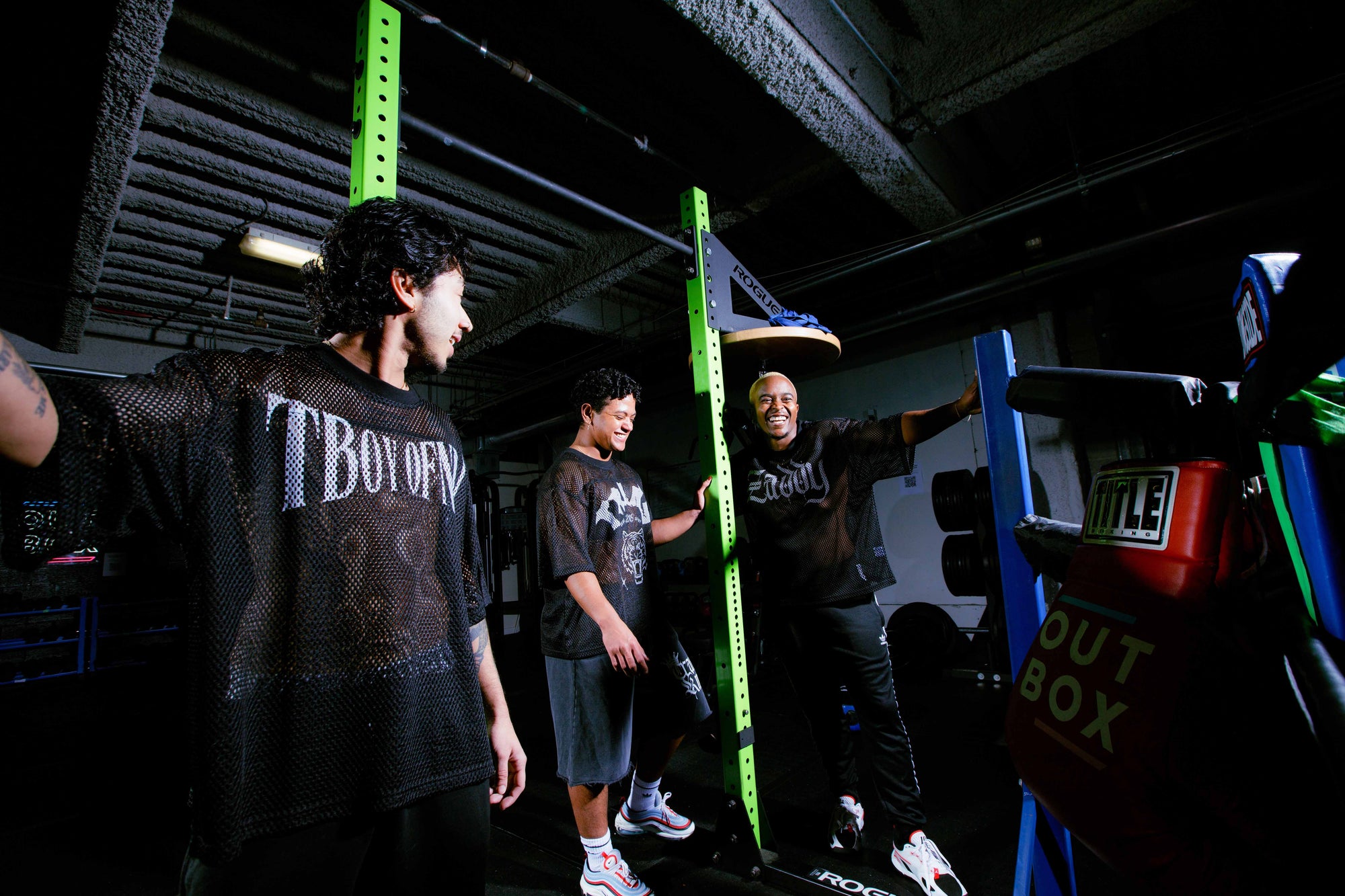 Three transmasc people in a gym setting with sports equipment, smiling and laughing.