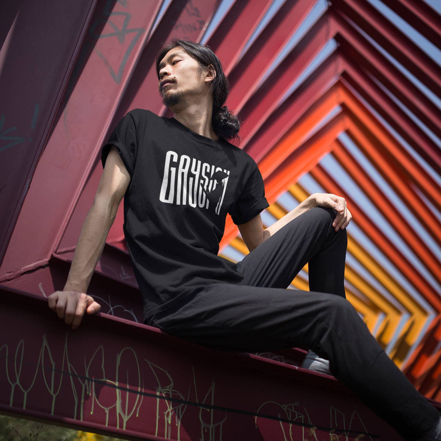 Person wearing a black t-shirt with 'Gaysian' typography with negative space creating the side profile of a warrior, sitting against a colorful geometric wall