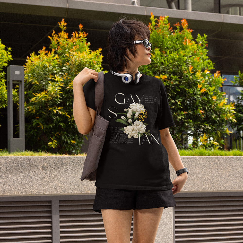 Person wearing a black t-shirt with floral design that says Gaysian in a subtle and stylish way with and shorts, standing outdoors.