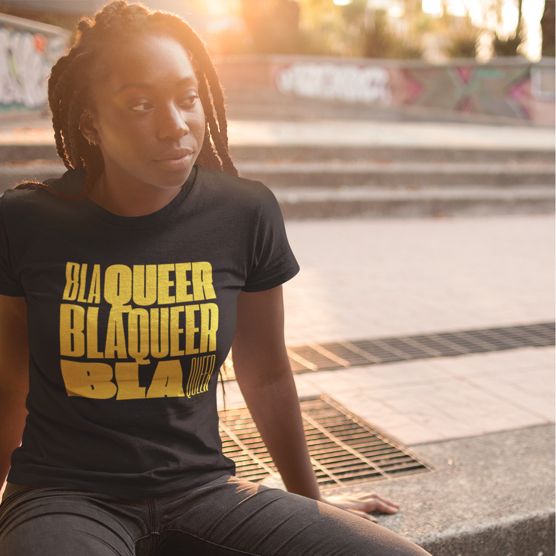 Black Queer Woman wearing a black t-shirt with 'BLAQUEER' text, sitting on a concrete surface.